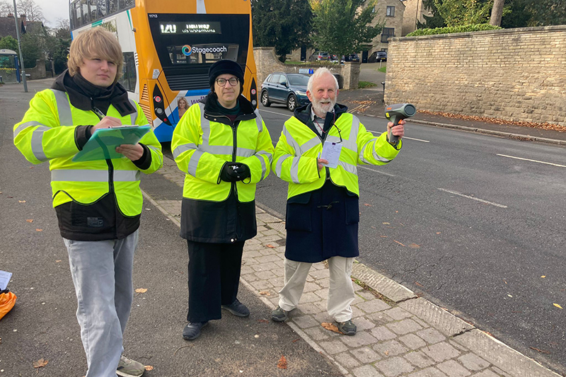 Three people in hi vis jackets by a roadside. One is holding a speed gun and another is holding a clipboard